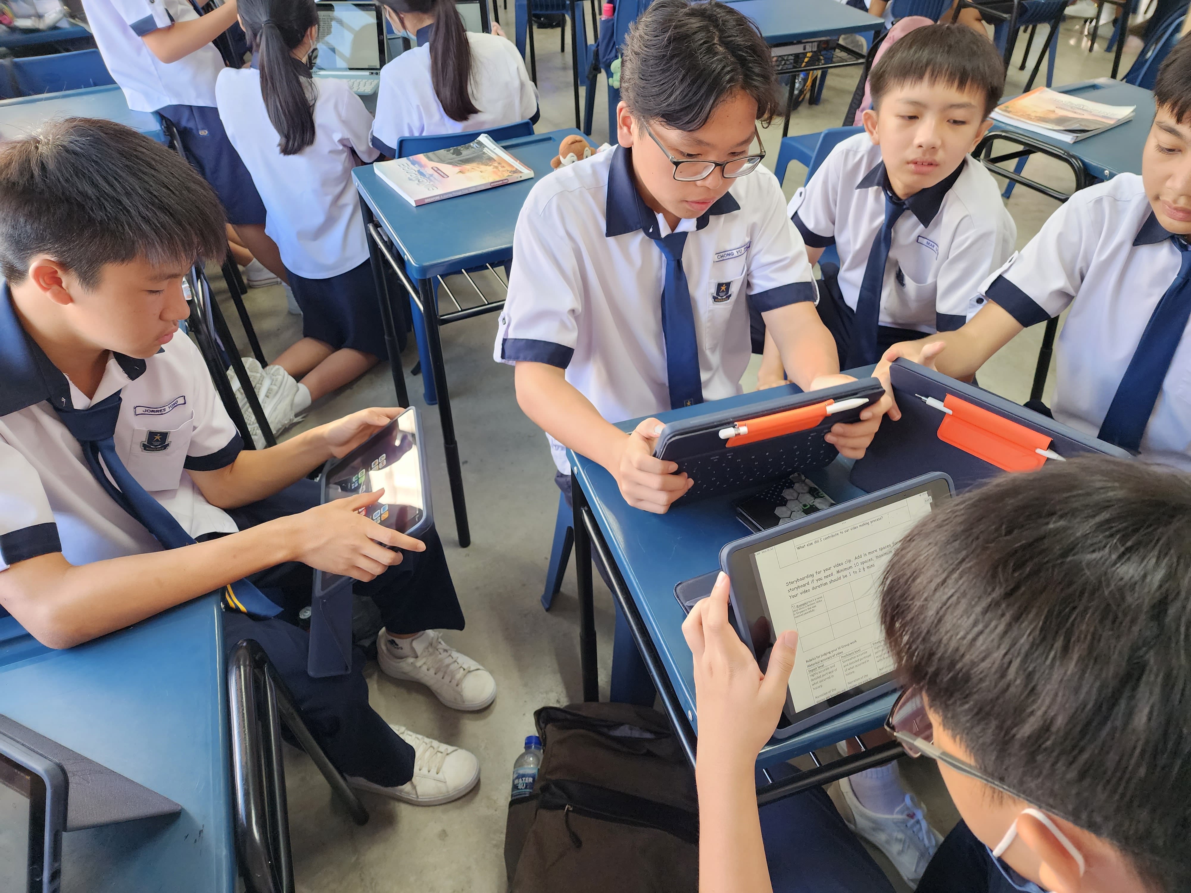 Students in white and navy uniforms sit at blue desks in a classroom, each using a tablet device.
